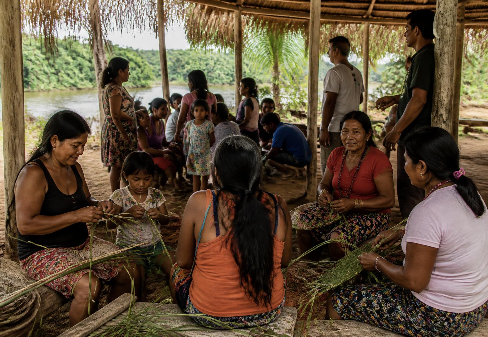 Moradoras e crianças da comunidade reunidas sob uma maloca de palha à beira do rio, trançando fibras vegetais durante uma atividade coletiva.